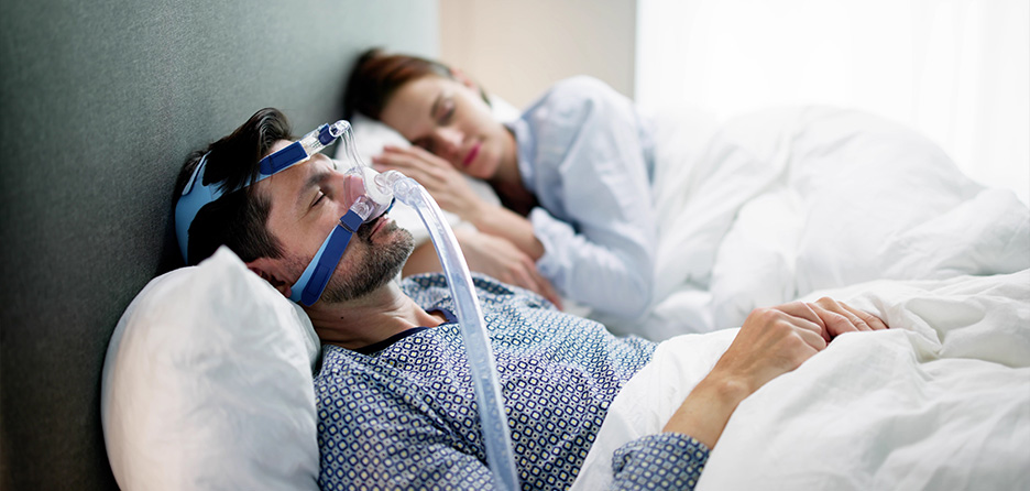 Man wearing a CPAP mask while sleeping to treat his sleep apnea, with his partner asleep beside him.